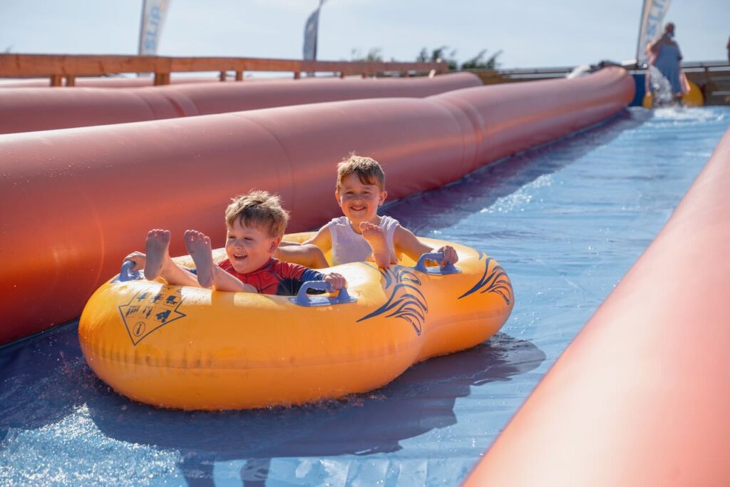 Two boys smiling in a yellow inflatable ring on the giant slide at Cornwall Slip and Slide, Atlantic Reach Resort near Newquay